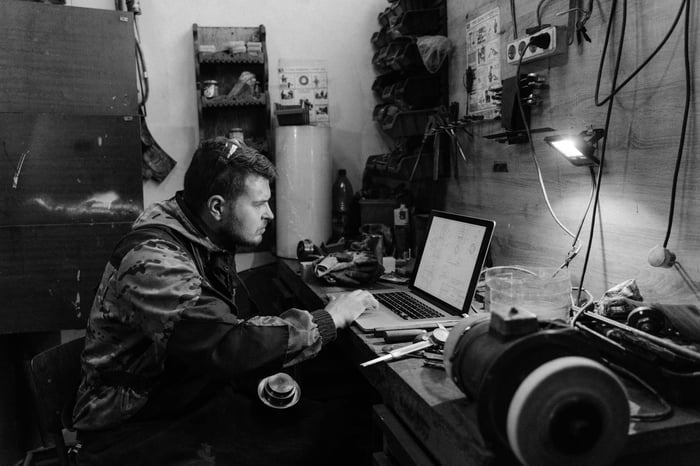 Black and white photo of a craftsman working on a laptop in a workshop setting.